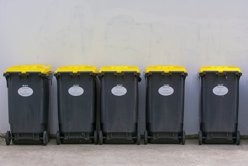 A row of five large, black wheeled rubbish bins with yellow lids is positioned on a concrete surface against a plain, light-colored wall. Each bin has a white oval label with black text and barcodes,