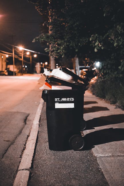 A row of five large, black wheeled rubbish bins with yellow lids is positioned on a concrete surface against a plain, light-colored wall. Each bin has a white oval label with black text and barcodes,