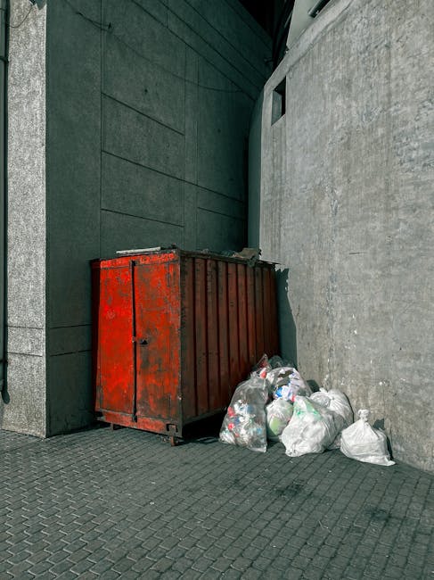 A large, weathered red metal dumpster positioned against a dark green wall and a light grey concrete wall in an urban outdoor setting. The dumpster has visible rust patches and a corrugated metal surf
