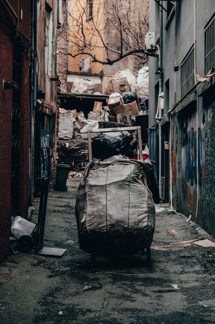 A narrow urban alleyway filled with various waste and rubbish, including black and white plastic bags, cardboard boxes, and loose debris, piled up against a makeshift barrier at the back of the alley.