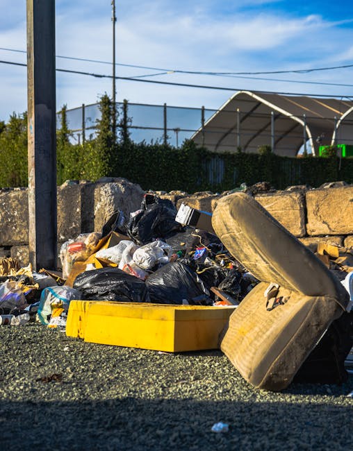 A narrow urban alleyway filled with various waste and rubbish, including black and white plastic bags, cardboard boxes, and loose debris, piled up against a makeshift barrier at the back of the alley.